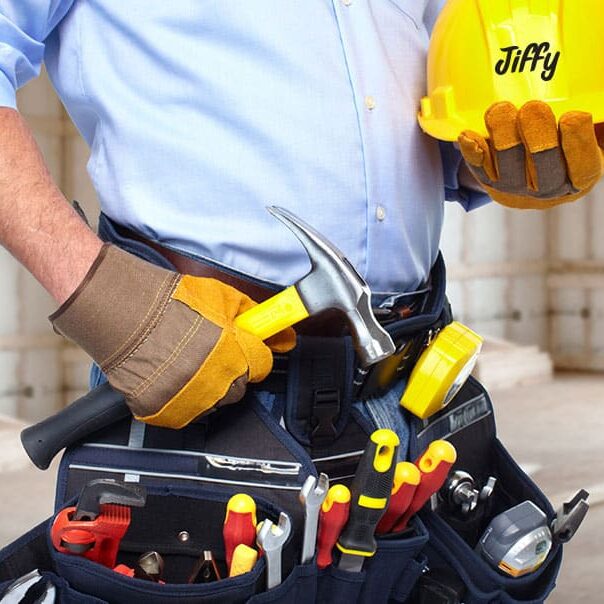 A construction worker wearing gloves and a tool belt holds a hammer and a yellow hard hat labeled Jiffy. The tool belt is filled with various tools, including screwdrivers, pliers, and a measuring tape.
